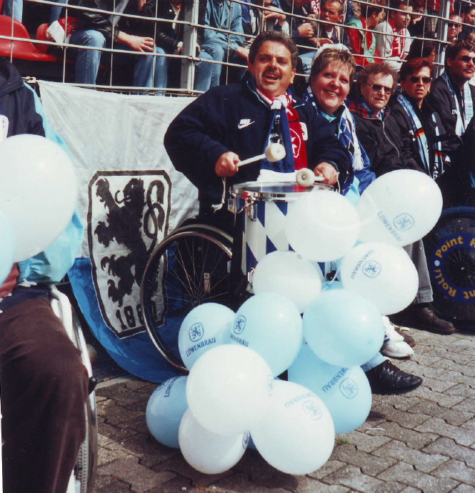 Auf Zuschauerplätzen im Stadion sind Fans im Rollstuhl mit einer Trommel auf den Beinen. Im Hintergrund die Vereinsflagge. Im Vordergrund viele Luftballons.