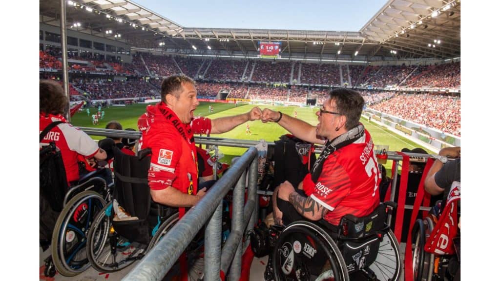 Zwei im Rollstuhlsitzende und Trikottragende SC-Fans. Beide befinden sich im neuen SC Stadion auf der Tribüne. Im Hintergrund der Spielfeld und die gegenüberliegende Tribüne zu erkennen.