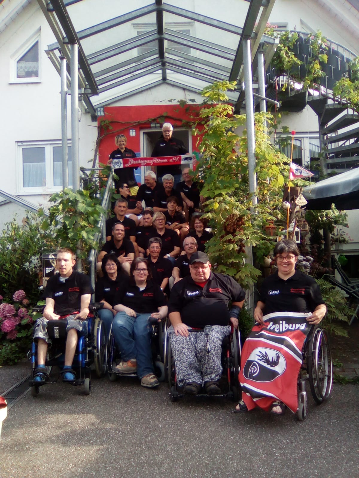 Gruppenfoto der Vereinsmitglieder vor einem Hauseingang. Mitglieder sitzen auf der Treppen und Rollstuhlfahrende davor.
