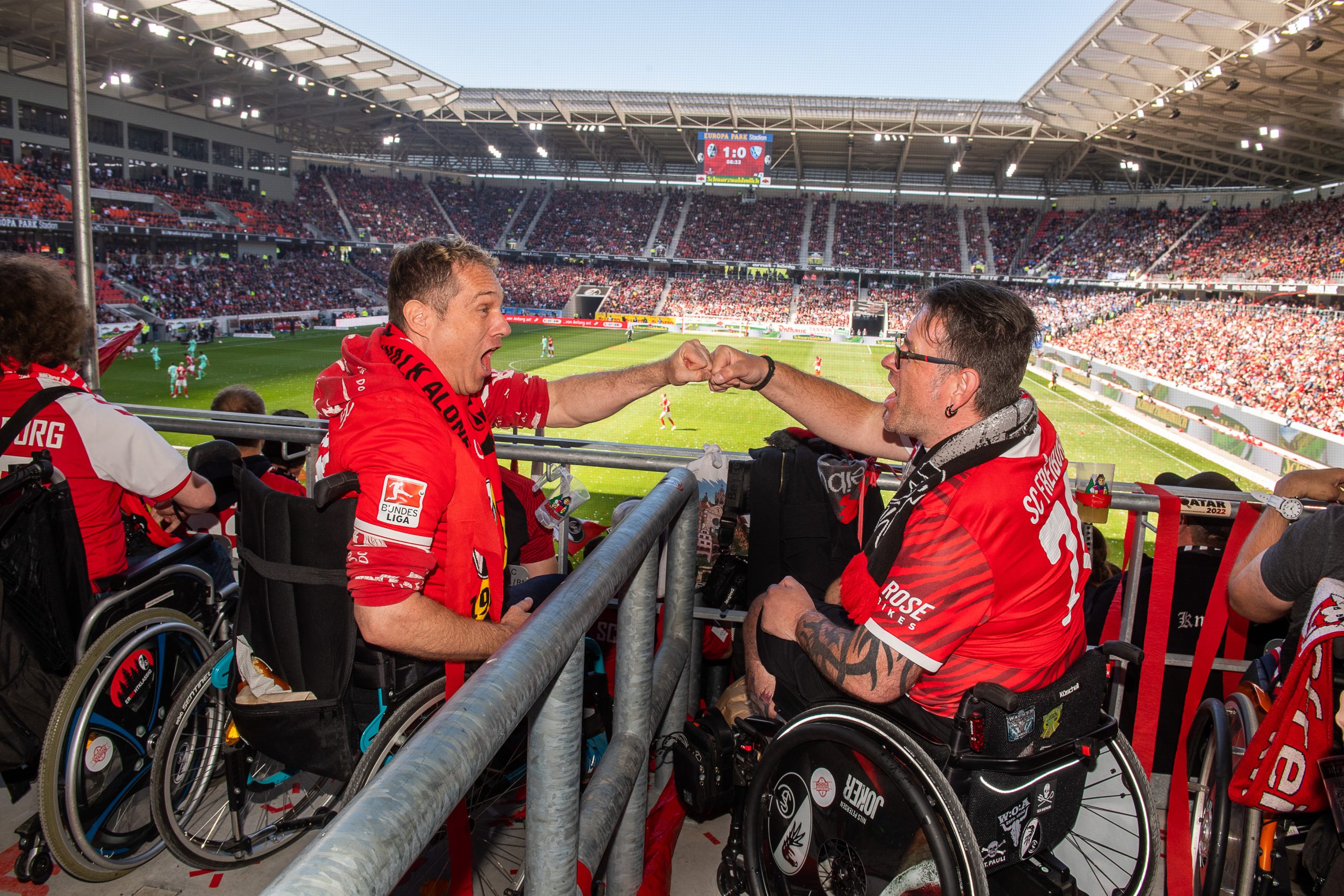 Zwei im Rollstuhlsitzende und Trikottragende SC-Fans. Beide befinden sich im neuen SC Stadion auf der Tribüne. Im Hintergrund der Spielfeld und die gegenüberliegende Tribüne zu erkennen.
