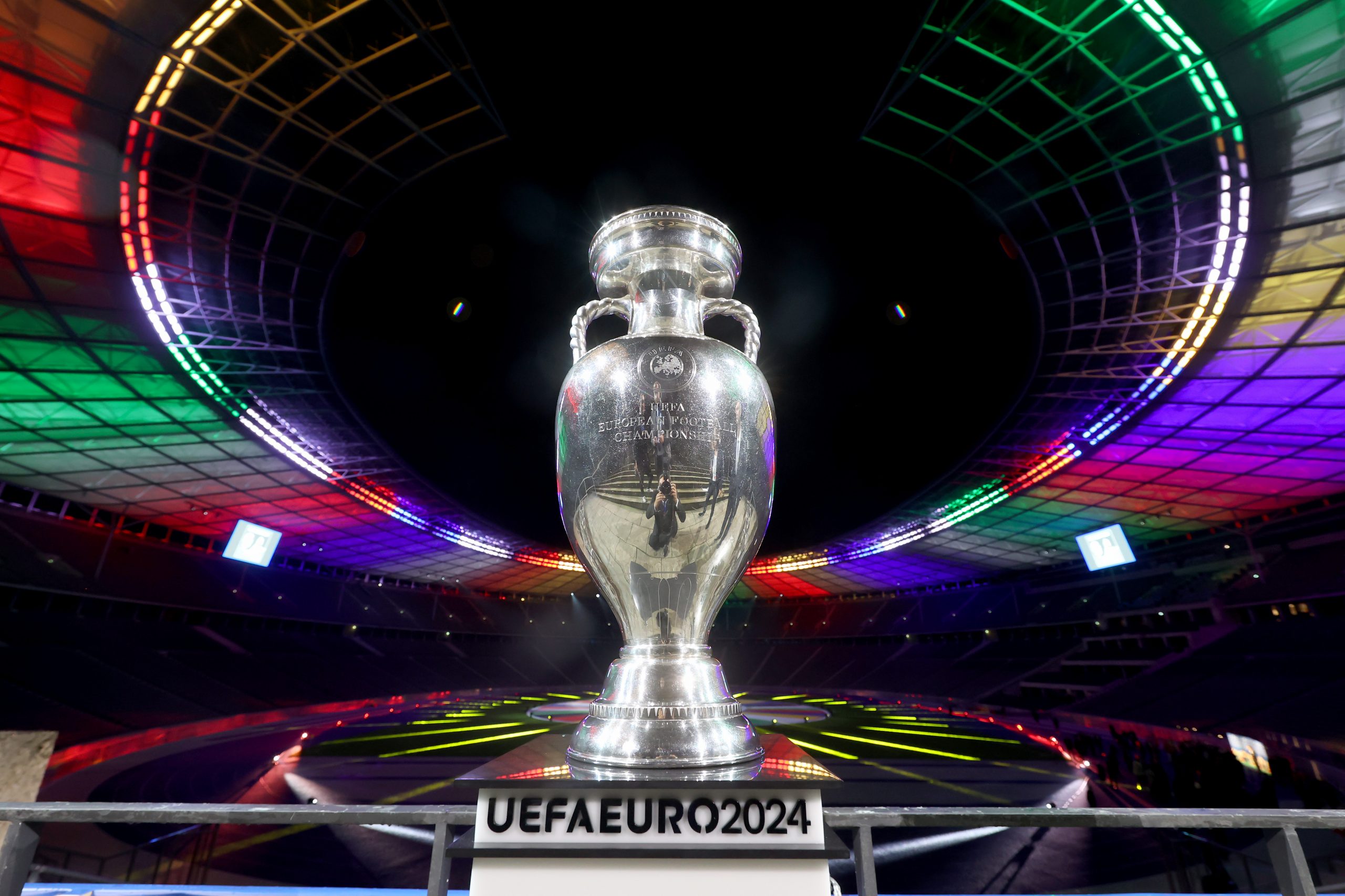 Im Hintergrund ist ein buntbeleuchtetes Stadion bei Nacht zu sehen. Sowohl das Dach als auch der Fußballplatz werden beleuchtet. Im Vordergrund ist der silberne UEFA Euro 2024 Pokal mittig auf dem Bild auf einer Säule zu erkennen. Der innen Raum des Stadions ist ganz dunkel.