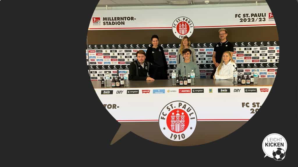 Leicht Kicken-Gruppenfoto im Pressekonferenzraum des FC St. Pauli. Sechs Personen formieren sich hinter den drei Mikrofonen.